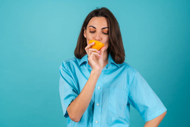 Young woman in a blue shirt on the background holds an orange, poses cheerfully, in high spirits, laughs smiles - Fotoğraf, Görsel