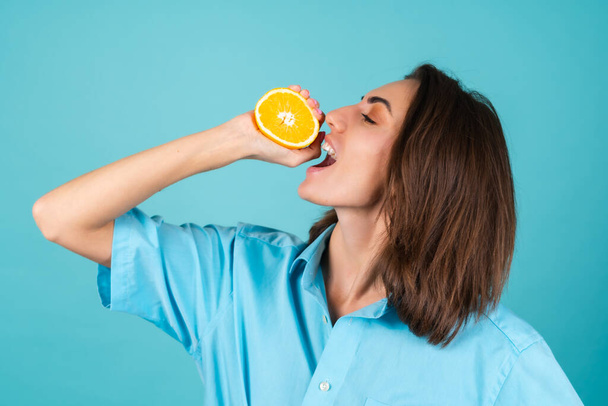 Arka planda mavi gömlekli genç bir kadın elinde neşeyle poz veren bir portakal tutuyor, morali yüksek, ağzına bir portakal sıkıyor. - Fotoğraf, Görsel