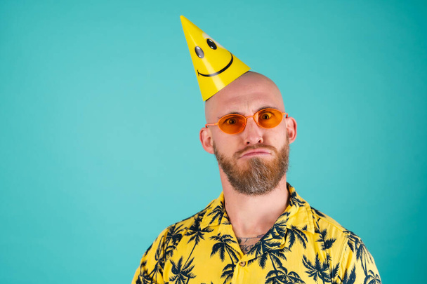 Γενειοφόρος άνδρας σε πουκάμισο με αποτύπωμα παλάμης σε τυρκουάζ φόντο γιορτάζει τα γενέθλια, χαζολογώντας με καπέλα για τις διακοπές - Φωτογραφία, εικόνα
