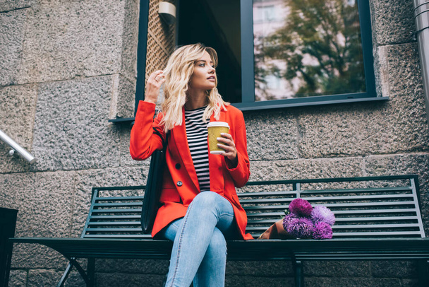 Rozważanie turystki z filiżanką napoju kofeinowego na wynos siedzi na ławce z bukietem fioletowych wiosennych kwiatów, przemyślana hipsterka z kawą na wynos myśląc o kwiatowym prezencie - Zdjęcie, obraz