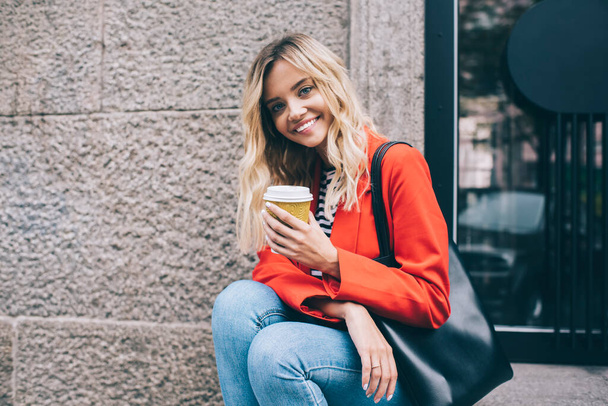 Half length portrait of happy Caucasian fashionista holding caffeine beverage and smiling at camera during photo session at street, cheerful hipster girl with coffee to go posing in travel city - Фото, изображение
