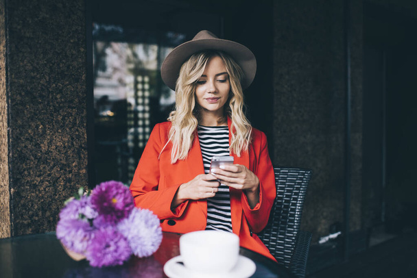 Millennial female blogger in trendy hat using mobile application for online chatting in social media, Caucaisan hipster girl with violet bouquet connecting to 4g wireless for browsing web content - Foto, Bild