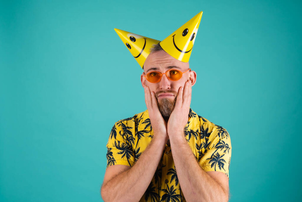 Γενειοφόρος άντρας σε πουκάμισο με σχέδιο φοίνικα σε τυρκουάζ φόντο, στα γενέθλιά του, με καπάκια για τις γιορτές, δυστυχισμένος και μόνος - Φωτογραφία, εικόνα