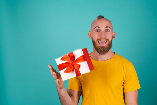 Un hombre barbudo con una camiseta naranja sobre un fondo turquesa lanza una caja de regalo en un estado de ánimo eufórico, da alegría, sonríe agradablemente - Foto, Imagen