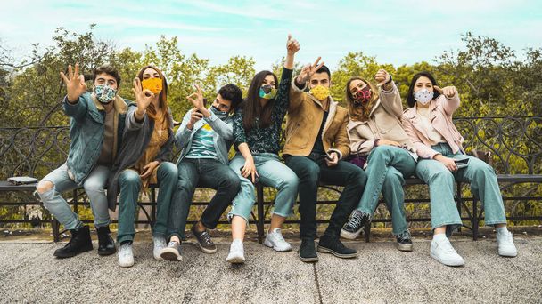 Glückliche junge Leute mit Gesichtsmaske sitzen auf einer Bank und amüsieren sich gemeinsam im Freien - neues normales Konzept. - Foto, Bild