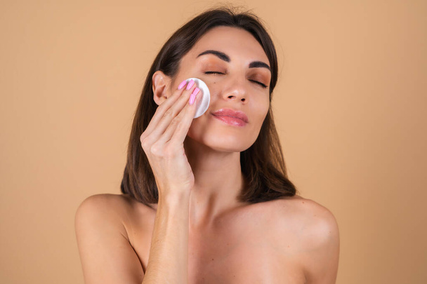 Close up portrait of a topless woman with bare shoulders on a beige background holding a sponge for cleaning her face, beauty portrait, taking care of her face skin - 写真・画像