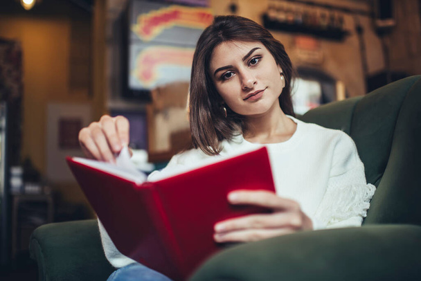 Focused woman student in casual clothes looking at camera while sitting in armchair reading text book while preparing for exam in cafe - Foto, imagen