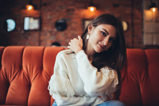 Glad female in casual clothes inclining head and touching hair sitting on soft sofa in light cafe while having break - Fotoğraf, Görsel