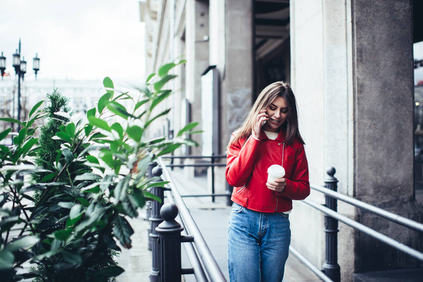 Smiling female with long hair wearing casual outfit walking near concrete building with takeaway coffee and having conversation on mobile phone - Photo, image