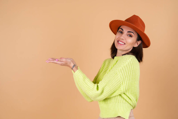 Модная стильная женщина во весь рост на бежевом фоне в брюках, свитере и шляпе, держа в руке пустое пространство, улыбаясь - Фото, изображение
