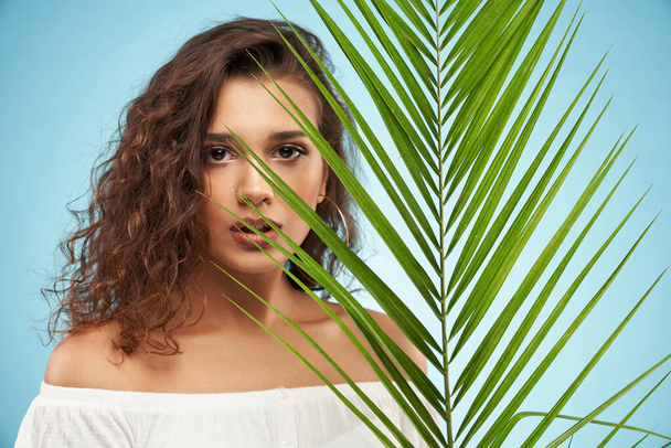 Pretty woman behind palm leaf, isolated on blue. - Foto, Imagem