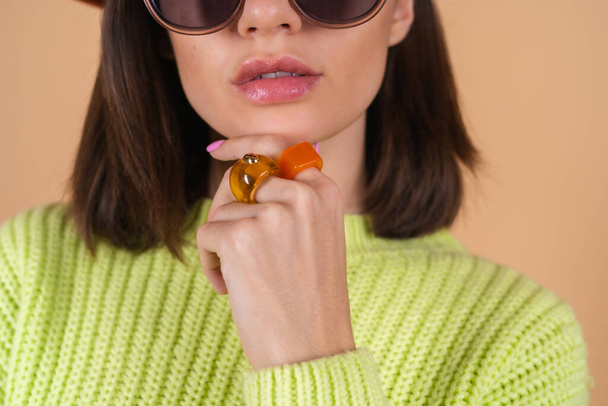 Модна стильна жінка на бежевому фоні в светрі і капелюсі і сонцезахисних окулярах позує, великі об'ємні кільця на пальцях
 - Фото, зображення