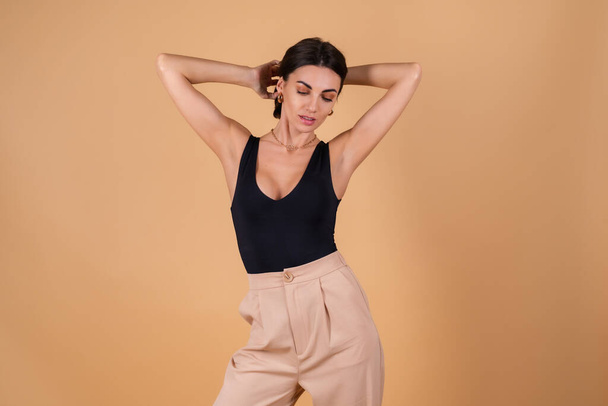 Fashionable portrait of a young woman on a beige background in a black bodysuit and trousers with gold cuts, a chain, a ring, posing - Foto, imagen