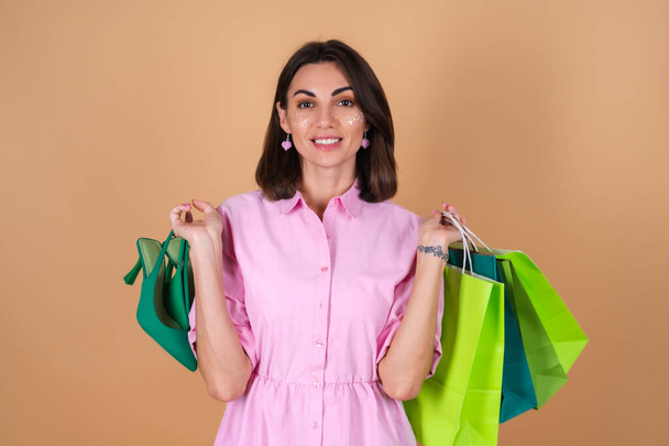 Portrait of a young woman in a pink dress on a beige background with glitter makeup holding shoes and shopping packages cute smiling - Фото, изображение