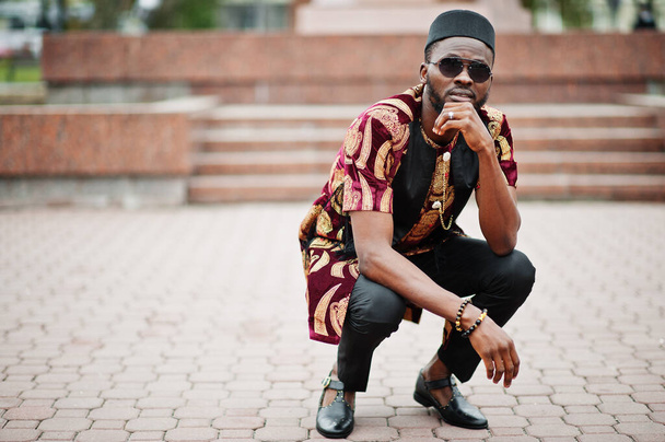 African stylish and handsome man in traditional outfit and black cap squatted outdoor. - Zdjęcie, obraz