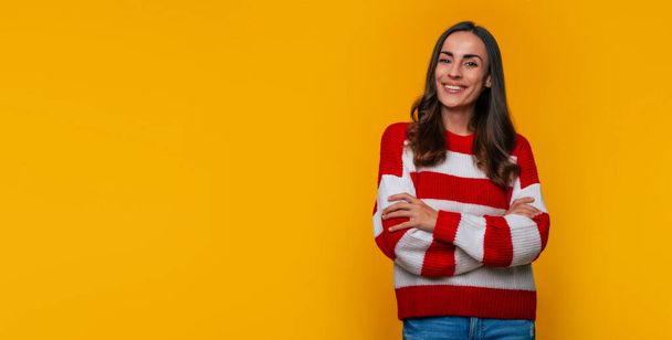 Banner photo of a cute smiling young woman in a sweater with crossed hands while she posing isolated on yellow background - Foto, Imagem