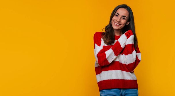Charming happy beautiful smiling young woman in striped sweater while she posing, looking on camera and isolated on yellow background - 写真・画像