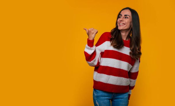 Happy and excited lovely smiling young woman in a sweater while she pointing away at advertising and isolated on yellow background - 写真・画像
