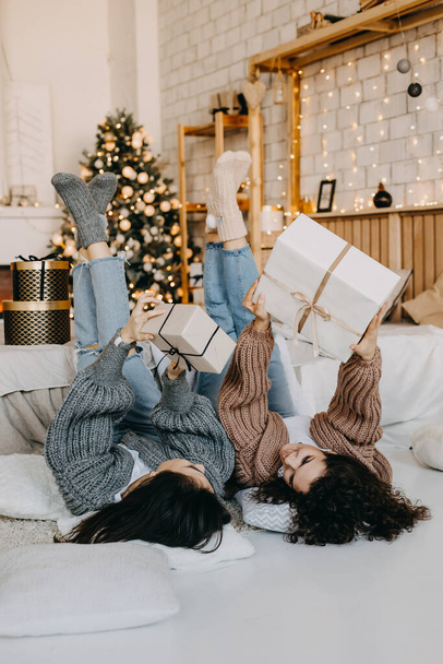 Two happy young women lying on the floor on christmas tree background, holding wrapped gift boxes. - Foto, imagen