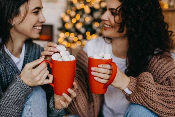 Two happy brunette women at home on christmas tree background, smiling, holding red cups with cocoa and marshmallows. - Photo, Image