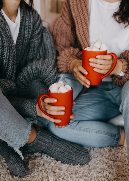 Closeup of two women wearing knitted sweaters and socks, at home, holding red cups with cocoa and marshmallows. - Fotó, kép
