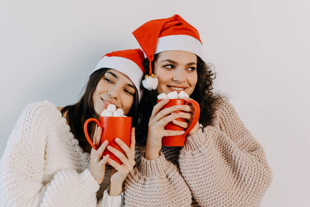 Deux jeunes femmes brunes heureuses portant un chapeau de Père Noël, souriantes, tenant des tasses rouges avec des guimauves. - Photo, image