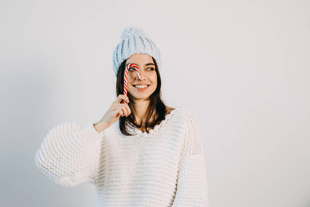 Cheerful young woman wearing a knitted beanie, smiling, holding a candy cane. - Photo, Image
