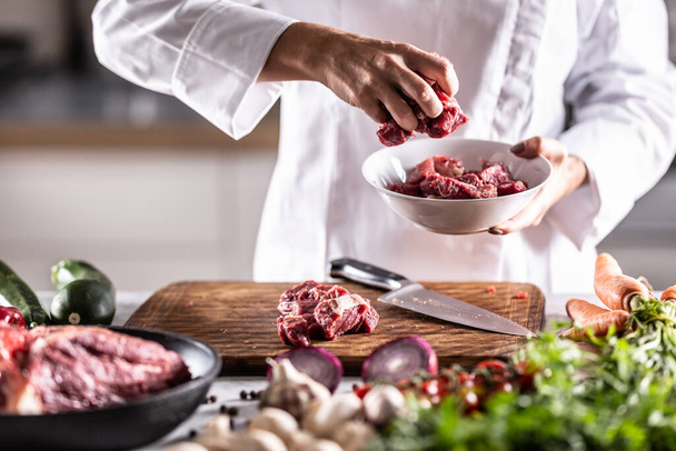 Detail of red meat chunks put into a white bowl on a meal preparation table full of ingedients. - Photo, image