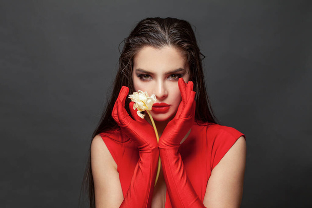 Pretty young sexy model female with makeup and dark hair in red dress and red silk gloves posing and holding white flower in black studio background - Photo, Image