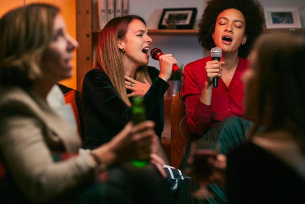 Ταλαντούχοι πολυφυλετικοί φίλοι κάθονται στο σπίτι και τραγουδούν ντουέτο στο σπίτι τη βραδιά καραόκε. - Φωτογραφία, εικόνα