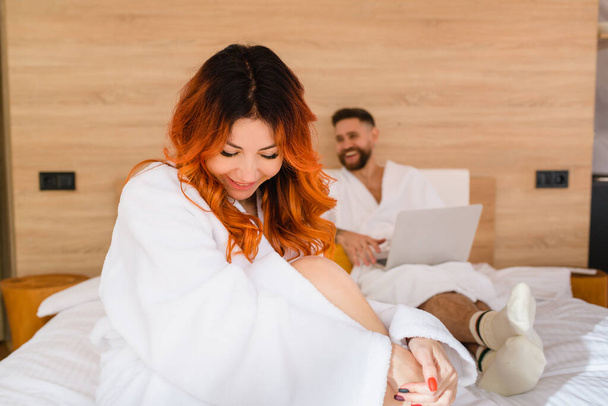 Porträt einer lächelnden attraktiven jungen Frau mit roten Haaren im weißen Bademantel, die auf dem Bett sitzt. - Foto, Bild