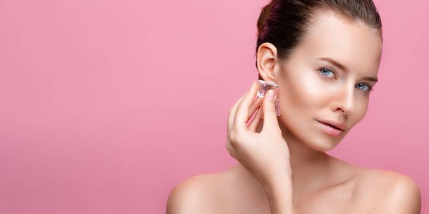 mooie brunette Kaukasische vrouw met perfecte huid houdt ijsblokje in de buurt gezicht op roze achtergrond - Foto, afbeelding