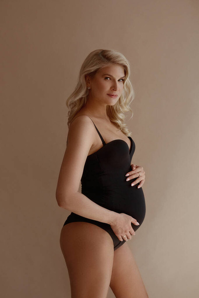Charming pregnant woman with blond hair in black bodysuits posing on the beige background - Foto, Imagem