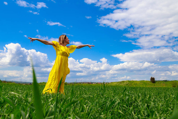 Young woman in a yellow dress outdoors in a green field. Hands raised to the sky as a symbol of happiness and freedom. - Foto, immagini