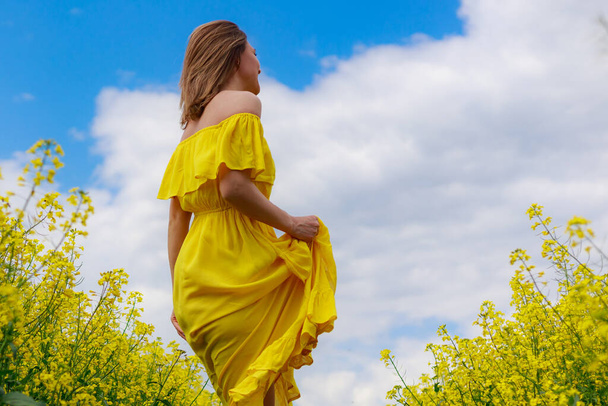 Young pretty woman in yellow dress on lightning cheerful yellow background of blooming rapeseed field. Good mood concept - Photo, Image