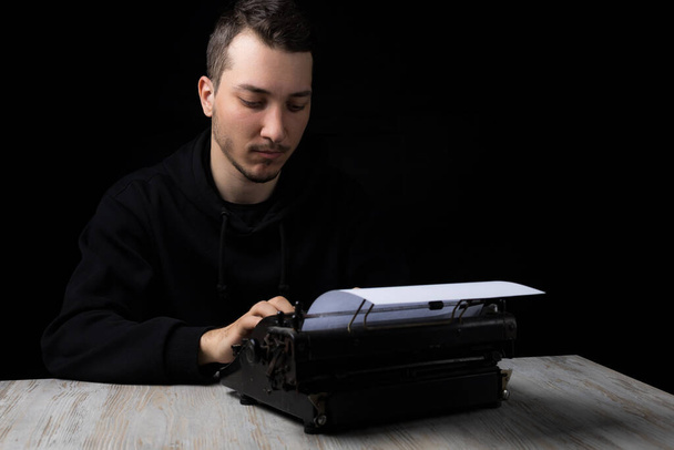 Mladý pohledný muž v černé mikině je psaní na psacím stroji, na černém pozadí v low key - Fotografie, Obrázek