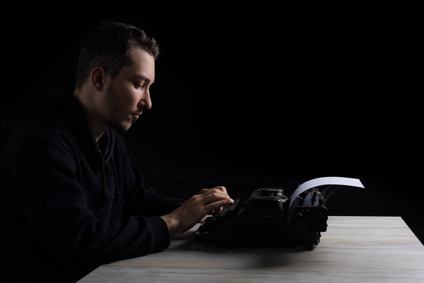 Junger gutaussehender Mann in schwarzem Kapuzenpulli tippt auf einer Schreibmaschine, auf schwarzem Hintergrund in leisem Ton - Foto, Bild