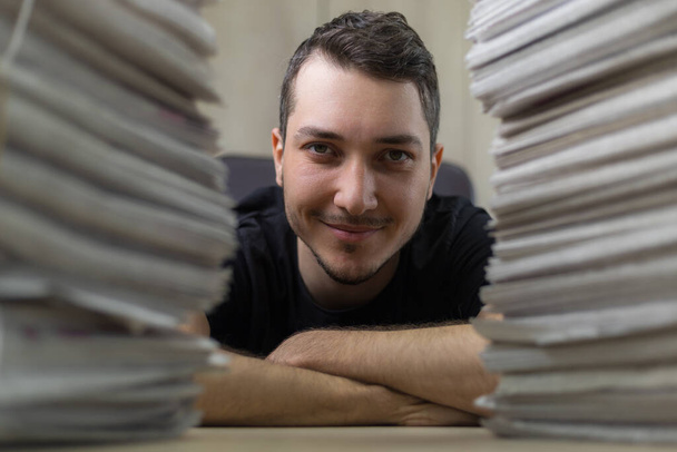 Close up portrait of a young journalist and a stacks of newspapers in a blurred foreground. - Photo, Image