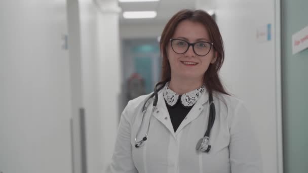 Fejlövés portré mosolygó, millenniumi női orvosról, aki orvosi egyenruhát és sztetoszkópot visel, miközben a kamerát nézi egy modern kórház folyosóján. Egészségügyi koncepció, egészségbiztosítás - Felvétel, videó