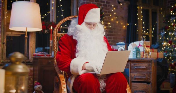 Close up of happy senior man in Santa Clause costume typing at the laptop while sitting at home. Holidays concept - Fotoğraf, Görsel