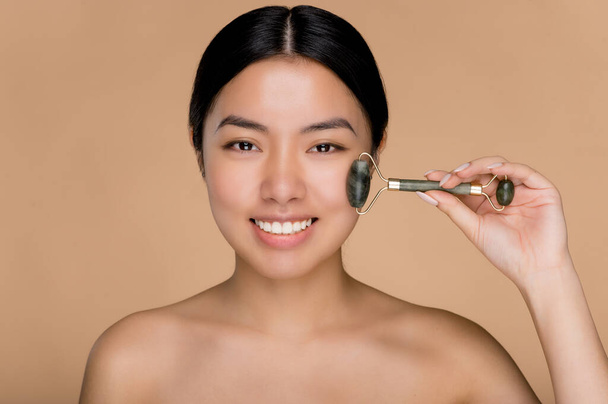 Close up photo of charming beautiful young Asian woman with naked shoulders, using derma roller for massage her facial skin. Photo of a smiling woman on isolated beige background - Fotó, kép
