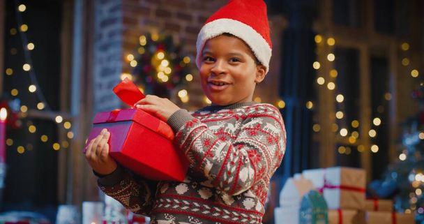 Little multiracial boy wearing Santa hat holding gift box with present while spending time at home with New Year decorations. Christmas spirit concept - Foto, immagini