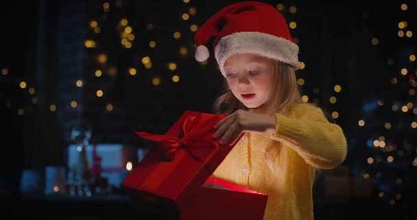 Little child girl in Santa hat opening gift box and wondering of the gift with Christmas Tree in the background - Foto, imagen