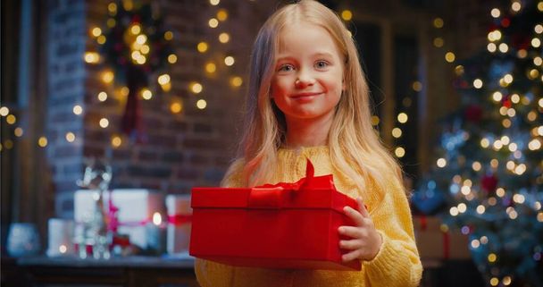 Little caucasian girl holding gift box with present while spending time at home with New Year decorations. Christmas spirit concept - Foto, immagini