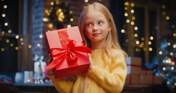 Little caucasian girl holding gift box with present while spending time at home with New Year decorations. Christmas spirit concept - Foto, imagen
