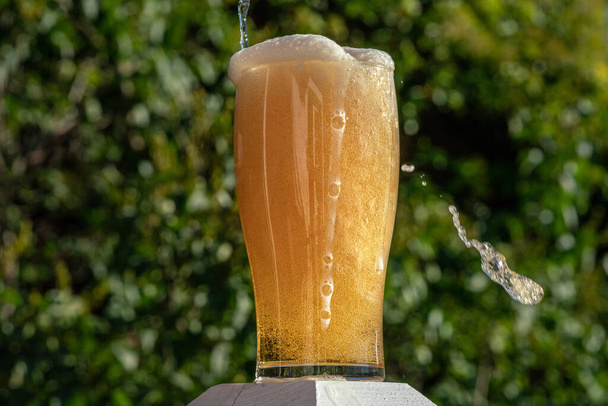 Ποτήρι μπύρας που χύνεται πάνω από την κορυφή με φυσαλίδες και αφρό που ρέει κάτω από το γυαλί, σε εξωτερικούς χώρους με έντονο ήλιο και πράσινο φόντο - Φωτογραφία, εικόνα