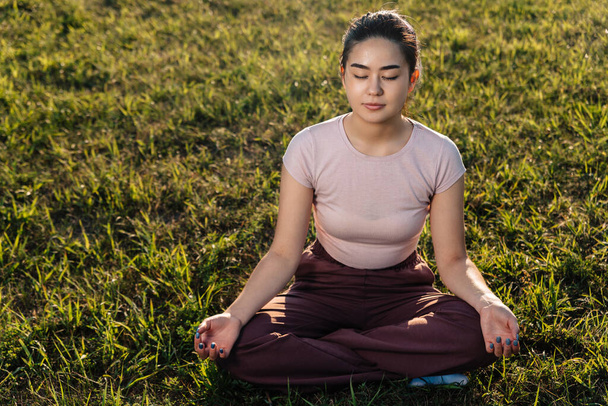 若いアジア系の女の子が芝生の上に座って瞑想している. - 写真・画像