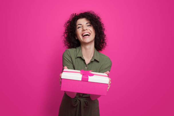 Smiling curly haired woman in dress posing while holding present box with gift ribbon bow isolated on pink background. Holiday gift surprise. Present birthday. - 写真・画像