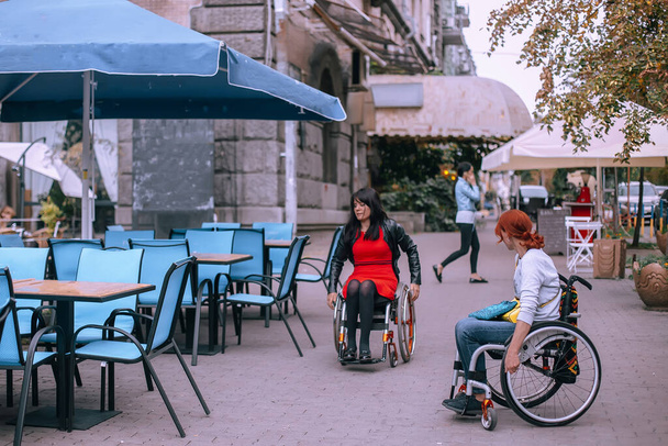 Dos chicas en sillas de ruedas en la ciudad cerca de la cafetería de la calle. Las mujeres con necesidades especiales caminan por las calles centrales de Kiev y se relajan en los restaurantes. Enfoque selectivo. - Foto, Imagen