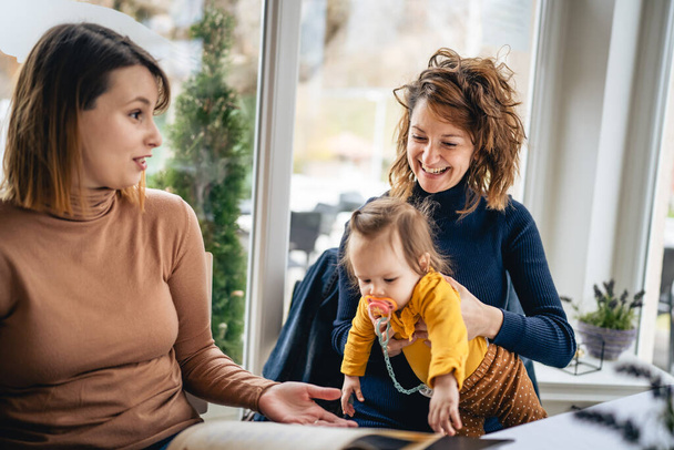 Woman hold child two female friends sitting at cafe or restaurant by the table holding small caucasian baby talking and smile happy real people friendship leisure concept in bright day selective focus - Photo, Image